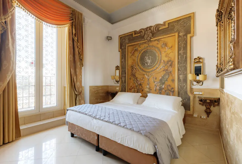 Guest room with a large gilded fresco headboard, tall curtained windows and a bed with grey quilted throw.
