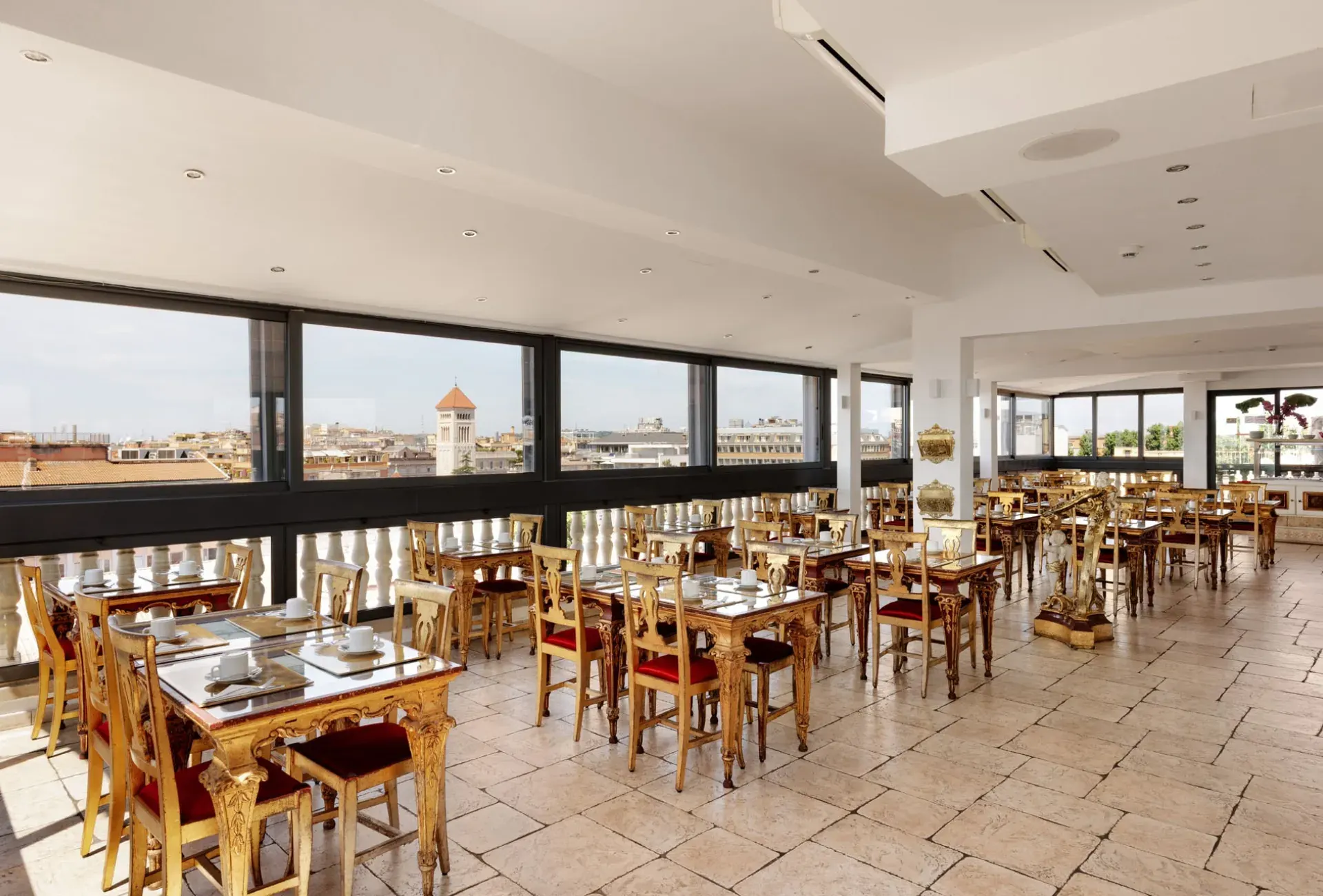 Bright rooftop restaurant with tables and gilded chairs under a long row of windows overlooking the rooftops of Rome.